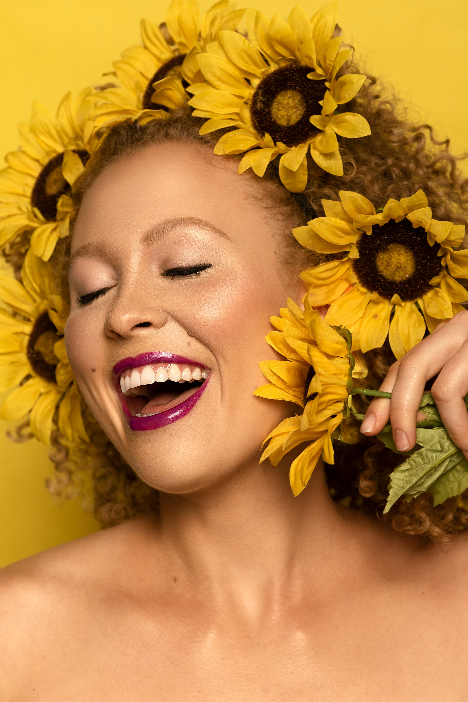 Female model smiling big with sunflowers covering her hair, eyes closed, closeup shot, featuring red lipstick by Utah photographer Christopher Loke