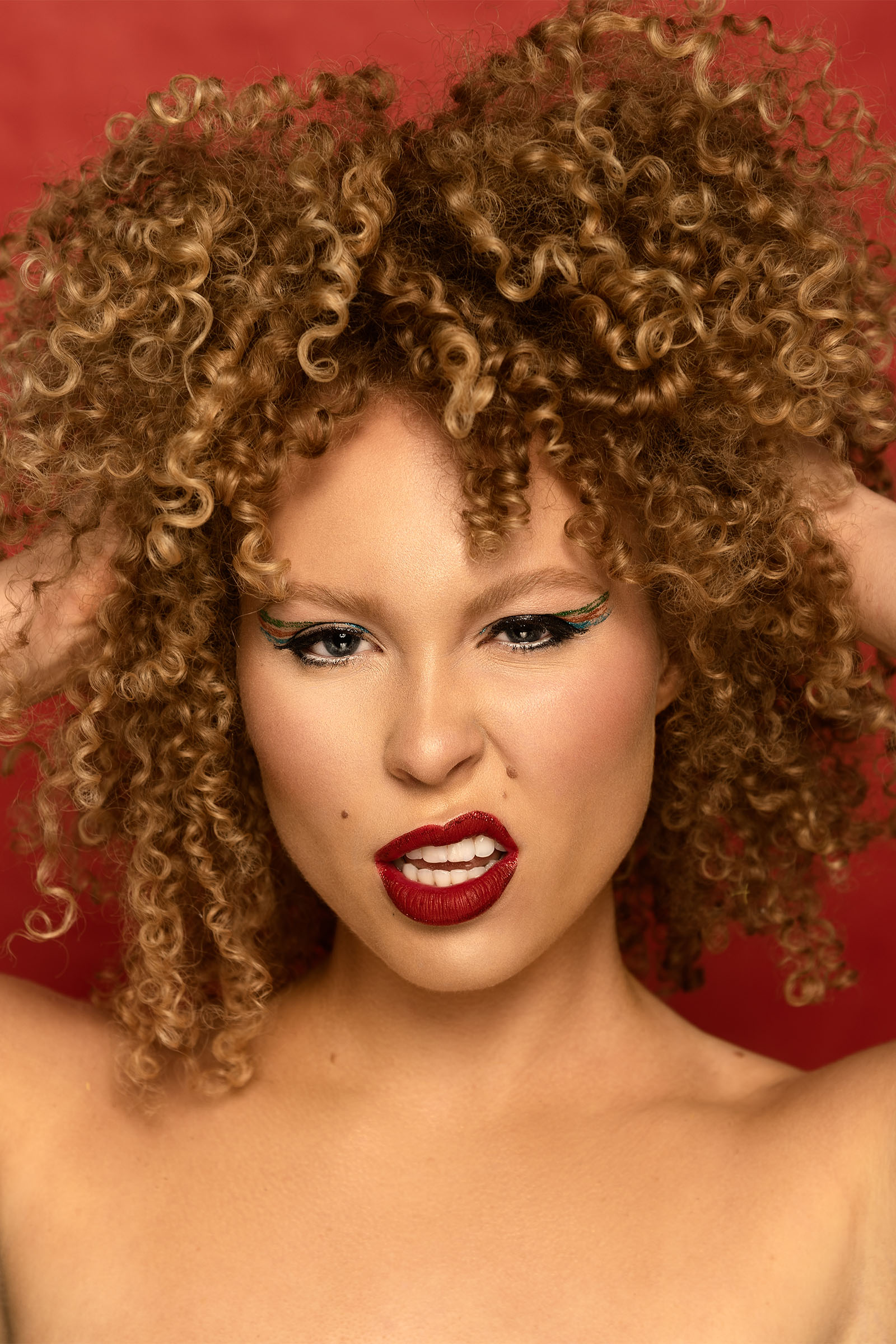Closeup beauty shot of female model featuring beautiful makeup, curly hair, hands on her hair, tousled, with a fierce but seduction expression by Utah photographer Christopher Loke